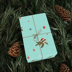 May include: A rectangular gift box wrapped in light blue paper featuring a repeating pattern of sea turtles wearing red and white Santa hats, red ornaments, and white snowflakes, tied with thin brown twine string across the center.