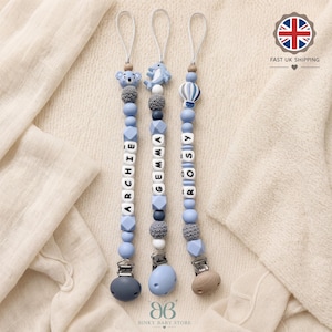 May include: Three personalised dummy clips featuring blue, grey, and white silicone beads, crochet beads, and letter blocks spelling ARCHIE, GEMMA, and ROSY. Each clip includes a unique silicone charm—a koala, a dolphin, and a hot air balloon—attached to a metal clasp.