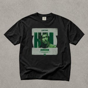 Pode incluir: Uma t-shirt preta de manga curta com uma impressão gráfica de um homem numa moldura de tom verde. O texto diz LANTERNS, JORDAN e o juramento: IN BRIGHTEST DAY, IN BLACKEST NIGHT, NO EVIL SHALL ESCAPE MY SIGHT.