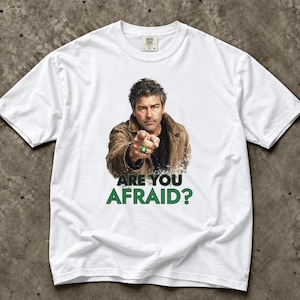 Może przedstawiać: Biały t-shirt z krótkim rękawem z nadrukiem graficznym przedstawiającym mężczyznę w brązowej kurtce, wskazującego palcem z zielonym pierścieniem. Pod obrazkiem znajduje się napis ARE YOU AFRAID? pogrubionymi, zielonymi wielkimi literami.