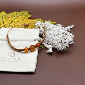 May include: A brown cord bracelet featuring three round, translucent orange carnelian beads. The bracelet is positioned on a beige fabric pouch with the printed text ALUNA Rooted in love, accompanied by a yellow maple leaf and white lichen.