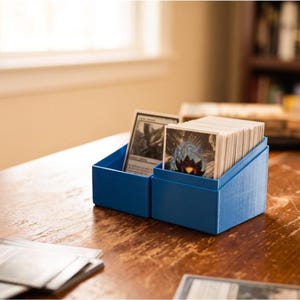 Può includere: Una scatola porta carte in plastica blu con parte superiore inclinata, contenente un mazzo di carte collezionabili. La scatola presenta due scomparti separati, uno contenente una singola carta e l'altro una pila di carte con illustrazioni.