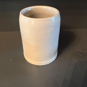 May include: A cylindrical ceramic mug with a smooth, light beige glaze and a slightly tapered shape. The vessel features a simple, unadorned exterior with a subtle rim at the base and a wide, open top, showcasing a clean and functional design.