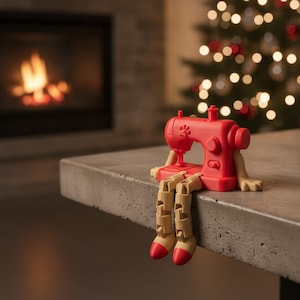 May include: A red sewing machine figurine with articulated beige limbs and red-tipped feet. The machine features a small paw print icon on its side and is designed to sit on the edge of a flat surface with its legs dangling over the side.