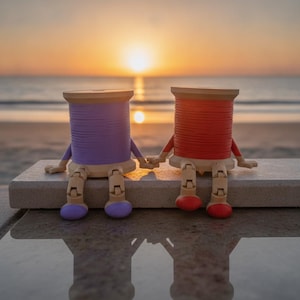 May include: Two anthropomorphic spools of thread, one purple and one red, featuring articulated wooden arms and legs. The figures are sitting side-by-side, holding hands, with their feet dangling down. The spools have a cylindrical shape.