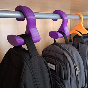 May include: Two purple and one orange plastic S-shaped hooks hanging from a silver metal closet rod. Each hook supports a black backpack by its top handle. The backpacks feature zippered compartments and a small white label with the word Name.
