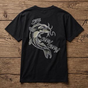 Op de afbeelding: Een zwart t-shirt met korte mouwen en een grafische illustratie van een meerval in grijstinten en wit. De tekst "HERE KITTY KITTY!" is in een gestileerd wit lettertype rondom de vis gedrukt, die opvallende snorharen en vinnen heeft.