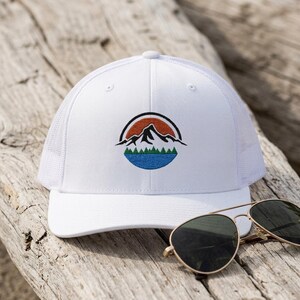 May include: A white trucker hat featuring an embroidered circular patch on the front. The design depicts a black mountain silhouette against an orange sun, with a green forest line and a blue lake below. The hat has a mesh back and a solid front.