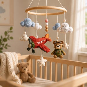 May include: A nursery mobile featuring a circular wooden frame with hanging crochet elements including a red airplane, a brown teddy bear wearing aviator goggles, white and blue clouds, and white stars, all suspended by white strings.
