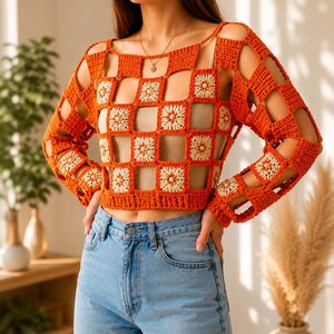 May include: A long-sleeved crochet top featuring a grid pattern of orange and cream-colored square motifs. Each cream square is accented with a small white bead in the center. The garment has an open-knit design and is paired with blue denim jeans.