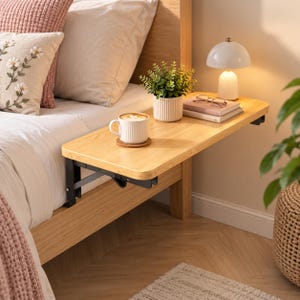 May include: A light-colored wooden bedside shelf attached to a bed frame with black metal folding brackets. The shelf holds a white ribbed mug on a coaster, a small potted green plant, a stack of two books, and a small white table lamp.