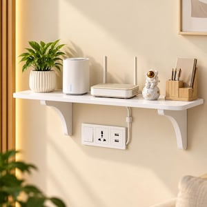Puede incluir: Un estante flotante blanco montado en la pared con dos soportes curvos, que sostiene una planta verde en una maceta blanca acanalada, un router cilíndrico blanco, un router inalámbrico blanco, una pequeña figura de astronauta y un organizador de madera.