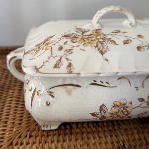 May include: A rectangular ceramic tureen with a lid, featuring a cream-colored base with brown floral patterns and gold-painted accents. The piece has a visible crackled glaze finish, two side handles, and a small handle on the lid.