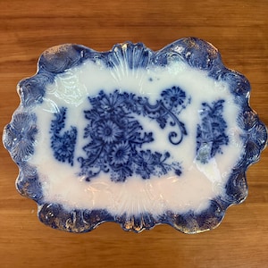 May include: An oval ceramic serving platter featuring a white center with a deep blue floral pattern. The scalloped edges are decorated with a dark blue and gold gilded border, showcasing intricate embossed shell-like details along the rim.
