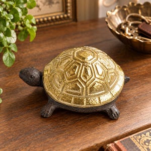 Può includere: Una statuetta a forma di tartaruga con un corpo in ghisa testurizzato color marrone scuro e un guscio dorato lucido e riflettente caratterizzato da un motivo geometrico esagonale. La tartaruga è raffigurata in posizione di cammino.