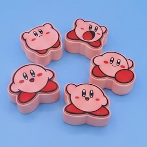 May include: Five pink erasers shaped like the character Kirby, each featuring a distinct facial expression including smiling, open-mouthed, and neutral faces, with red feet and a light pink body, arranged in a circular pattern on a light blue surface.