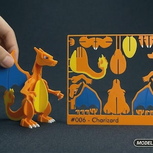 May include: An orange and blue Charizard model kit figure standing next to a flat plastic frame containing various orange and blue pre-cut parts. The frame is labeled #006 - Charizard and includes a small logo with the text MODEL KIT.
