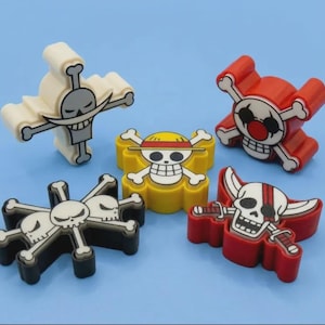 May include: Five plastic cookie cutters featuring various pirate skull emblems from One Piece. The set includes a white, red, yellow, black, and another red cutter, each shaped like a unique skull and crossbones design with distinct facial features.
