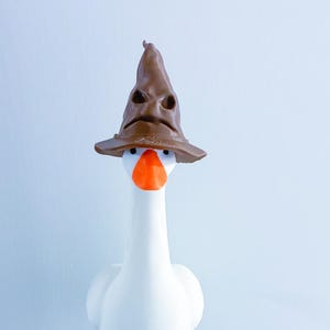 May include: A white goose figurine with an orange beak wearing a brown, weathered-looking pointed hat. The hat features deep, creased folds that resemble a face with eyes and a mouth, resting directly on top of the goose's head.