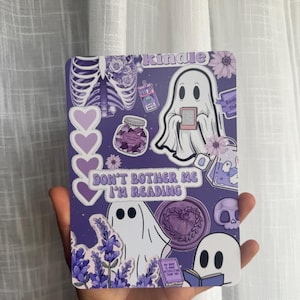 Può includere: Una custodia rettangolare viola per Kindle con un collage di illustrazioni di fantasmi bianchi, una gabbia toracica con fiori, rametti di lavanda, un teschio e cuori. Il testo riporta Kindle, Don't Bother Me I'm Reading e altre brevi frasi.