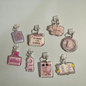 May include: A collection of eight clear acrylic charms with pink and purple designs featuring book-themed text and illustrations. Each charm includes a clear plastic attachment loop. The designs include phrases like Go away! I'm Reading, Bookish Girlie, and Emotional Support Kindle.