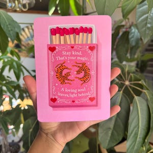 May include: A rectangular pink greeting card featuring an illustration of a matchbox with bright pink match heads. The box displays two orange tigers and the text: Stay kind. That's your magic. A loving soul leaves light behind. Red hearts decorate the border.