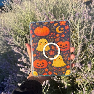 Op de afbeelding: Een doorzichtige tablethoes met een donkere achtergrond en een herhalend patroon van oranje pompoenen, lachende gele spookjes, rode bloemen en een maansikkel. Een witte, cirkelvormige magnetische ring is in het midden bevestigd.