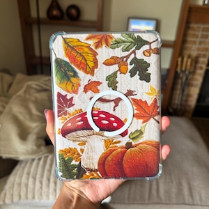 May include: A clear protective tablet case featuring a printed autumn-themed design with a white circular magnetic ring in the centre. The pattern includes colourful fall leaves, acorns, a red and white spotted mushroom, and an orange pumpkin.