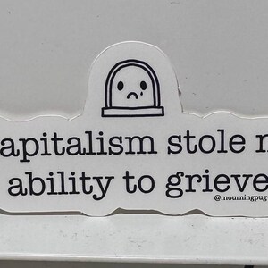 May include: A white sticker with black text that reads "Capitalism stole my ability to grieve." The sticker has a cartoon tombstone graphic with a sad face. The sticker also includes the text "@mourningpug."