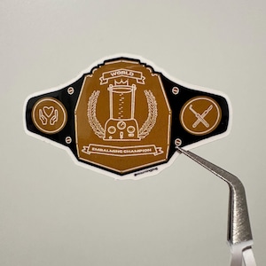 Può includere: Un adesivo a forma di cintura da campionato. L'adesivo ha una combinazione di colori marrone e nero con le parole "WORLD EMBALMING CHAMPION" e una grafica di frullatore. L'adesivo contiene anche il testo "@mourningpug".