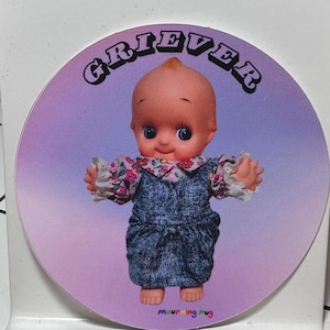 May include: A round sticker featuring a doll with a peach-colored head, blue eyes, and a denim overall outfit with floral sleeves. The word "GRIEVER" is arched above the doll. The text "mourning mug" is at the bottom.