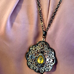 May include: A silver-toned filigree pendant necklace with a teardrop-shaped yellow gemstone. The pendant has an ornate, openwork design and hangs from a dark chain. The background is a soft, pinkish-purple fabric.