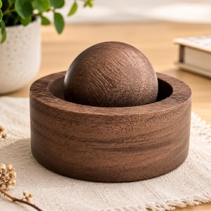 May include: A dark brown wooden bowl with a smooth, circular rim and a matching dark brown wooden sphere resting inside the center. The wood grain is visible across the surface of both the bowl and the sphere, showcasing a natural texture.
