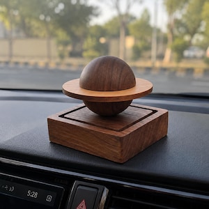 May include: A dark brown wooden sphere with a lighter brown wooden ring encircling its center, mounted on a square dark brown wooden base. The object is positioned on a black dashboard, featuring a smooth, polished finish on all wooden parts.