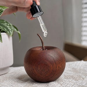 May include: A dark brown, apple-shaped wooden object with a smooth, rounded surface and a thin, curved stem. A clear glass dropper with a black cap is positioned above the apple, releasing a single droplet of clear liquid into the centre.