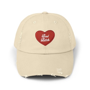 May include: A beige distressed baseball cap featuring a red embroidered heart patch on the front. Inside the heart, the text "Bad Bitch" is stitched in white cursive lettering. The cap has a worn-in look with frayed edges on the brim and crown.