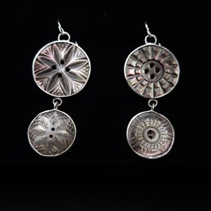 May include: A pair of drop earrings featuring two circular, dark, iridescent shell buttons each. The buttons are set in silver-toned metal frames and connected by small silver rings, hanging from silver-toned ear wires.
