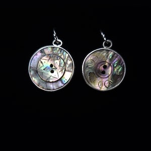 May include: Two circular iridescent abalone shell buttons with silver-toned metal rims and loops at the top. The left button features a star-like etched pattern, while the right button displays a series of overlapping circular engravings.