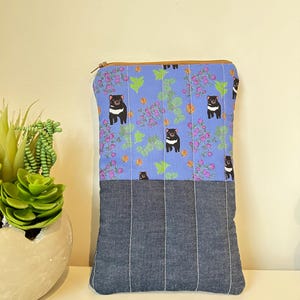 May include: A rectangular zippered pouch featuring a light blue upper section with a repeating pattern of Tasmanian devils, green leaves, and purple berries, and a dark gray lower section with vertical white stitching lines throughout the fabric.