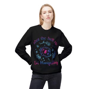 May include: A black long-sleeve sweatshirt featuring a graphic print of a skull surrounded by crystals, stars, and floral elements. The text "shut the hell up" and "I'm Manifesting" is printed in a gradient of blue, purple, and pink colors.