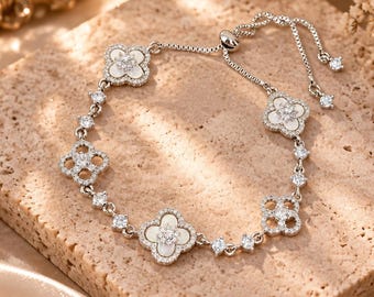 May include: A silver-toned adjustable bracelet featuring a series of four-leaf clover motifs. The design alternates between clover shapes inlaid with white material and openwork clover frames, all accented with small, clear, sparkling crystals.