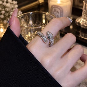 May include: A silver-toned open ring shaped like a snake, featuring a textured surface covered in small clear crystals and two small green stones for the eyes. The snake head and tail wrap around the finger, creating a bypass style design.