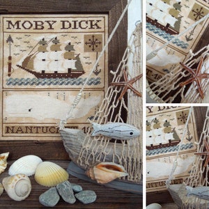 Puede incluir: Un bordado de punto de cruz enmarcado que representa un barco con el texto "MOBY DICK" y "NANTUCKET" sobre un fondo beige. El marco está rodeado por una cuerda marrón y una red con conchas y un pez de madera.