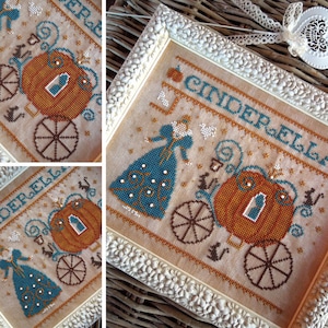 May include: A cross-stitch embroidery pattern of Cinderella in a pumpkin carriage with the text "Cinderella" above the carriage. The embroidery is framed in a white frame with a floral design.