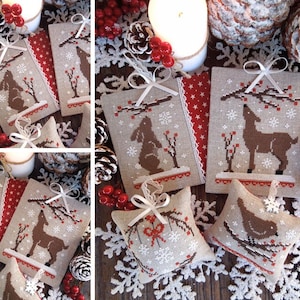 Puede incluir: Un conjunto de cinco adornos navideños hechos a mano con un ciervo, un conejo, un pájaro y un diseño de copo de nieve. Los adornos están hechos de tela blanca con bordados marrones y detalles rojos. Los adornos se muestran sobre una superficie de madera con piñas y bayas rojas.