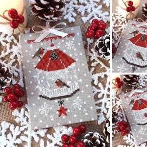 Può includere: Ornamento natalizio a punto croce raffigurante una casetta per uccelli con tetto rosso e due uccelli. La casetta è bianca con base marrone e una stella rossa. Lo sfondo è grigio chiaro con fiocchi di neve bianchi. Un nastro bianco in alto.