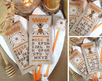 May include: Three cross-stitch bookmarks with a bee and alphabet design. The bookmarks are beige with orange and yellow accents. The alphabet is stitched in brown thread. The bee is stitched in brown, yellow, and black thread.