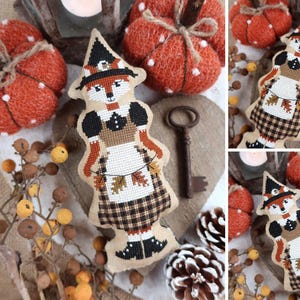 May include: A stuffed fabric ornament featuring a cross-stitched fox dressed as a witch, wearing a black hat, a brown bodice, and a plaid skirt. The fox holds a string of small autumn leaves. The ornament is light beige with detailed embroidery.
