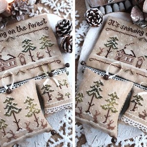 Puede incluir: Un sampler de punto de cruz con el texto "It's snowing on the forest" y una escena de invierno con una caba&ntilde;a, &aacute;rboles y nieve. El sampler est&aacute; enmarcado con un encaje blanco y tiene tres peque&ntilde;os adornos de tela marr&oacute;n con una escena de invierno similar.