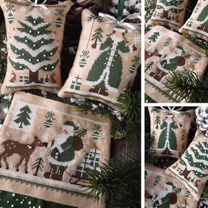 Puede incluir: Un conjunto de cinco adornos de punto de cruz con un árbol de Navidad, Papá Noel, un ciervo y dos mujeres con vestidos verdes. Los adornos están todos sobre un fondo beige con copos de nieve blancos y ramas de pino verdes.